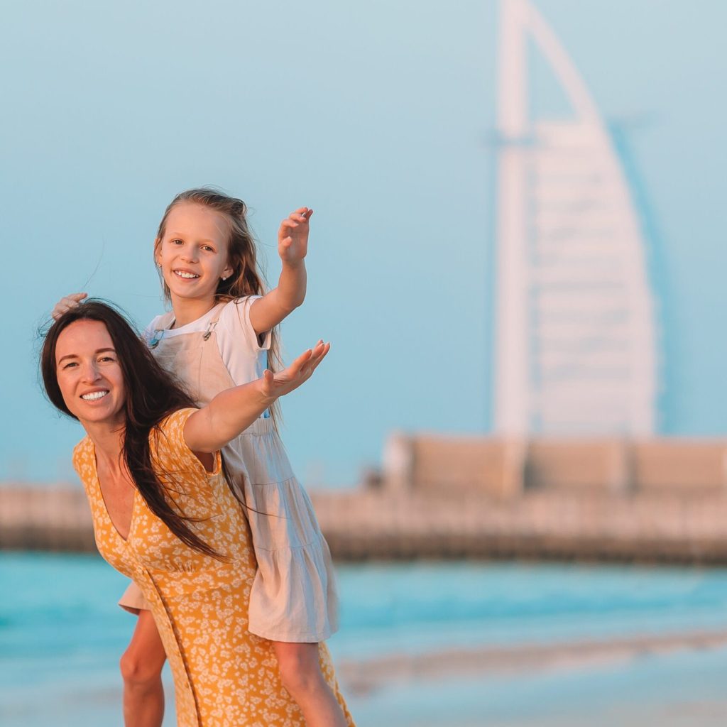 Young beautiful mother and her little daughter on the beach with Burj Al Arab on background in Dubai, UAE. United Arab Emirates famous tourist destination.