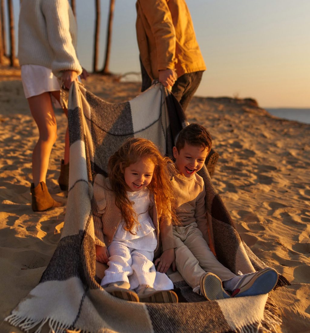 A young family, mom and dad, rolling two children on a warm blanket along the sandy beach by the sea at sunset, relax and have fun on weekends.