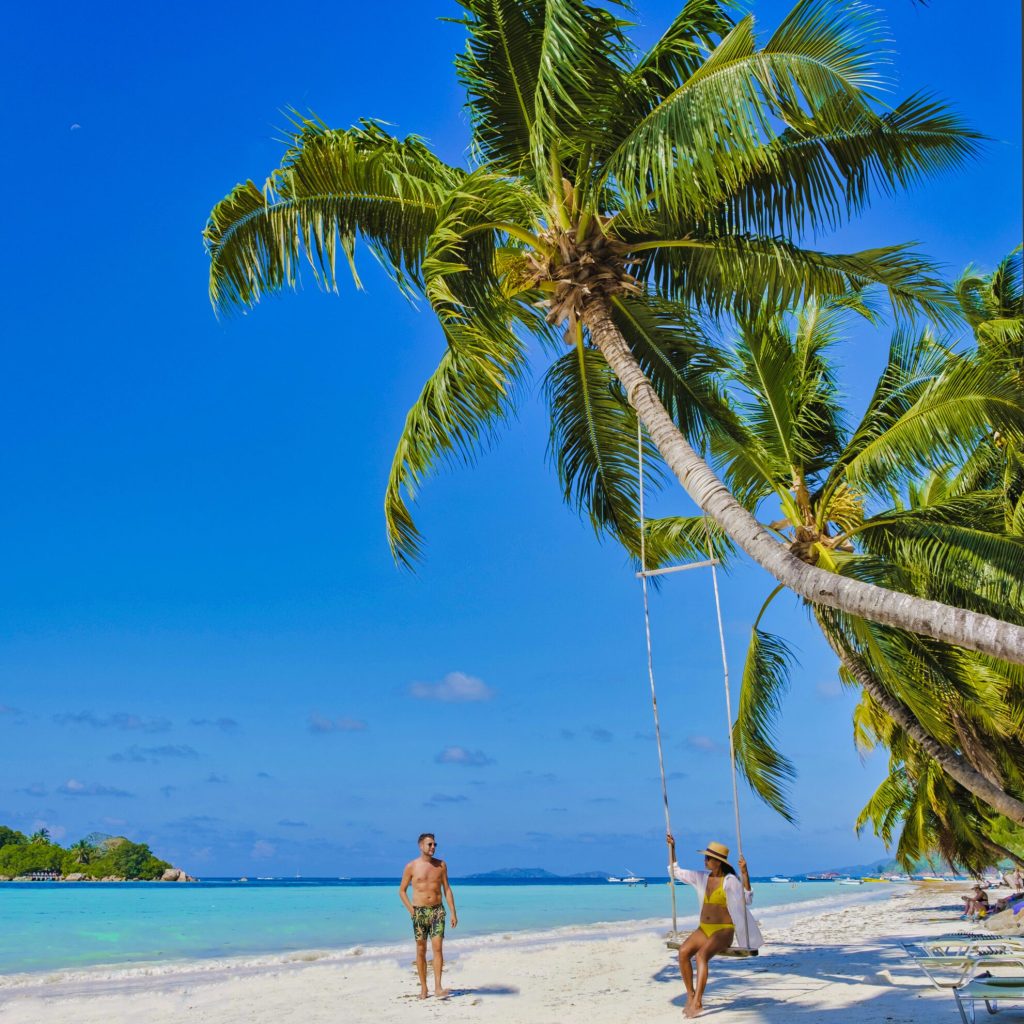 Praslin Seychelles tropical island with white beaches and palm trees, a couple of men and women in hammocks swing on the beach under a palm tree at Anse Volber Seychelles.